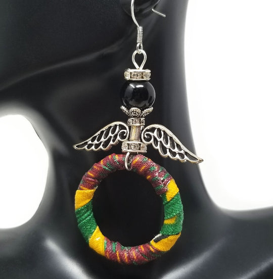 A pair of African-inspired angel earrings featuring hand-wrapped kente hoops in black, yellow, and green, with large silver angel wings and crystal accents.