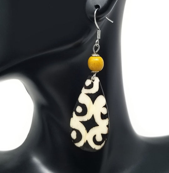 A pair of 'Dayo' Kenyan Batik Bone Earrings in yellow amber, featuring black and white patterned bone with a yellow amber bead, displayed on a mannequin ear.